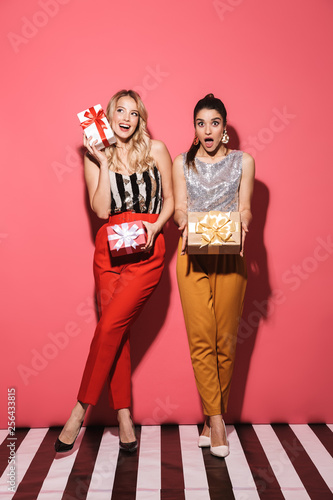Portrait of two excited girls 20s in stylish outfit on party holding gift boxes