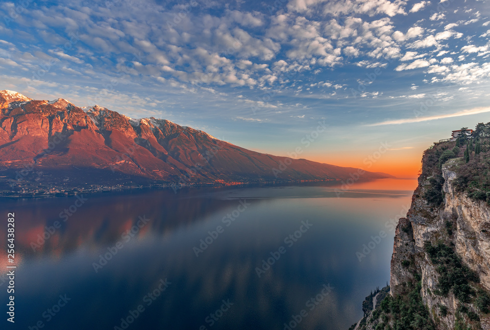 Stunning sunset on Lake Garda. View of the snowy Alps painted by the setting sun from the cliff of the city Tremosine. Winter period. Lombardy, Italy