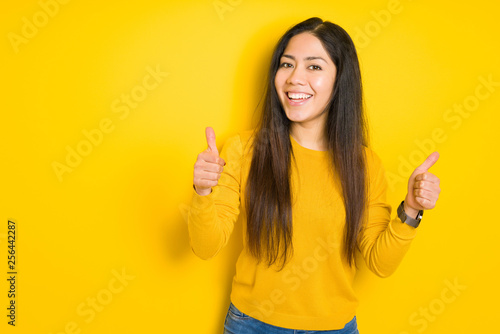 Beautiful brunette woman over yellow isolated background success sign doing positive gesture with hand, thumbs up smiling and happy. Looking at the camera with cheerful expression, winner gesture.