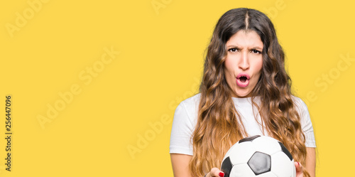 Young adult woman holding soccer football ball scared in shock with a surprise face, afraid and excited with fear expression © Krakenimages.com