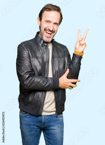 Middle age handsome man wearing black leather jacket smiling with happy face winking at the camera doing victory sign. Number two.