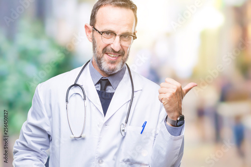 Middle age senior hoary doctor man wearing medical uniform isolated background smiling with happy face looking and pointing to the side with thumb up.