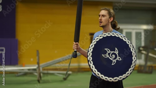 Young man with long hair instructs LARP foam combat class holding a shield and a sword inside a gym. 4k. photo