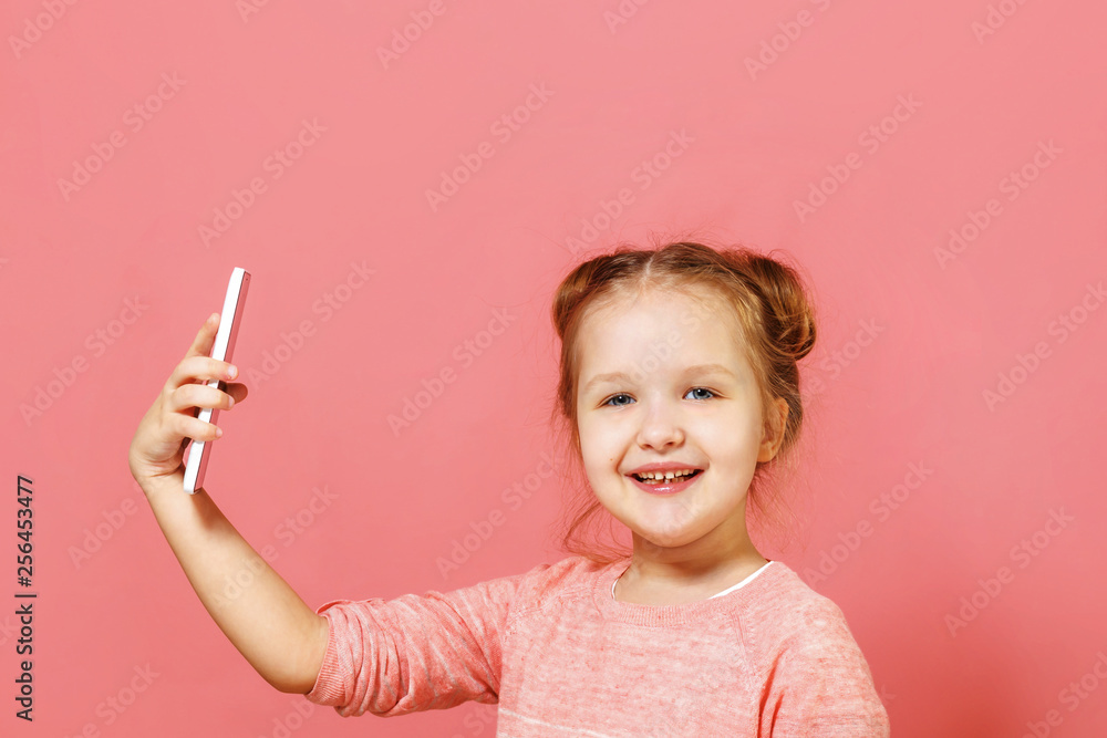Closeup portrait of a cute little girl with buns of hair on a pink background. The child holds the phone and takes a selfie Closeup portrait of a cute little girl with buns of hair on a pink background. The child holds the phone and takes a selfie