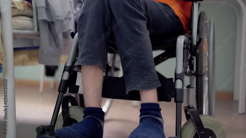 Close-up, low angle shot disabled young guy with spinal cord and leg problems (multiple sclerosis) puts feet on wheelchair footrest Close-up, low angle shot disabled young guy with spinal cord and leg problems (multiple sclerosis) puts feet on wheelchair footrest