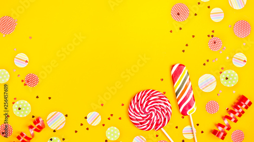 Two multicolored candies among paper circles of confetti, glitter and festive ribbons on a bright yellow background. Top view, flat lay, copy space