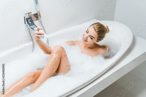 attractive and smiling woman taking bath with foam and taking selfie in bathroom