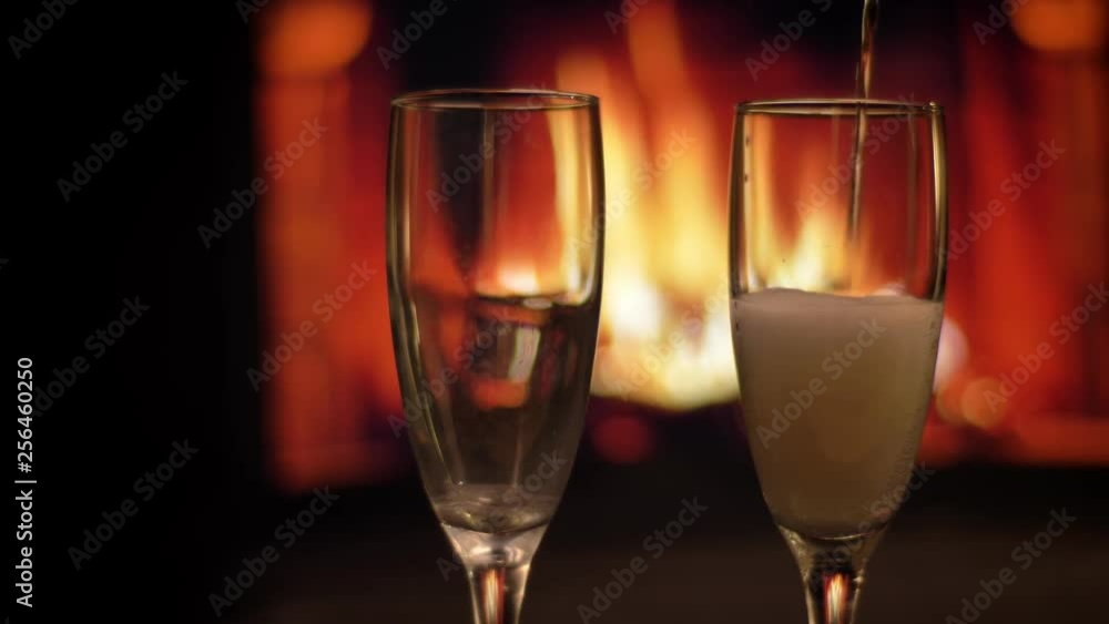 Closeup shoot of two empty glasses being poured a champagne into with cozy warm fireplace with flames on the background indoors in a dark apartment Closeup shoot of two empty glasses being poured a champagne into with cozy warm fireplace with flames on the background indoors in a dark apartment