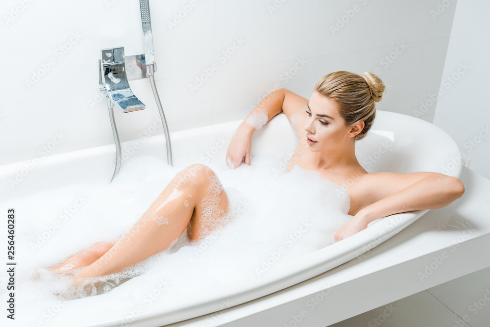 attractive and blonde woman taking bath with foam and looking away in  bathroom Stock Photo