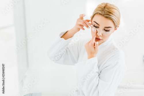 beautiful and blonde woman in white bathrobe applying eye patches in bathroom