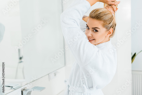 beautiful woman in white bathrobe playing with hair and looking at camera in bathroom