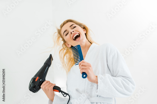 beautiful and smiling woman in white bathrobe singing and holding hairdryer, comb