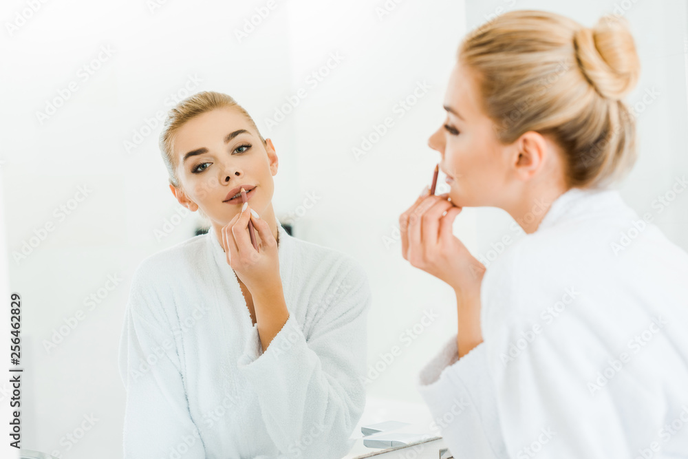 selective focus of beautiful and blonde woman in white bathrobe applying lip liner and looking at mirror selective focus of beautiful and blonde woman in white bathrobe applying lip liner and looking at mirror