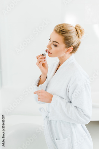 beautiful and blonde woman in white bathrobe applying lipstick in bathroom