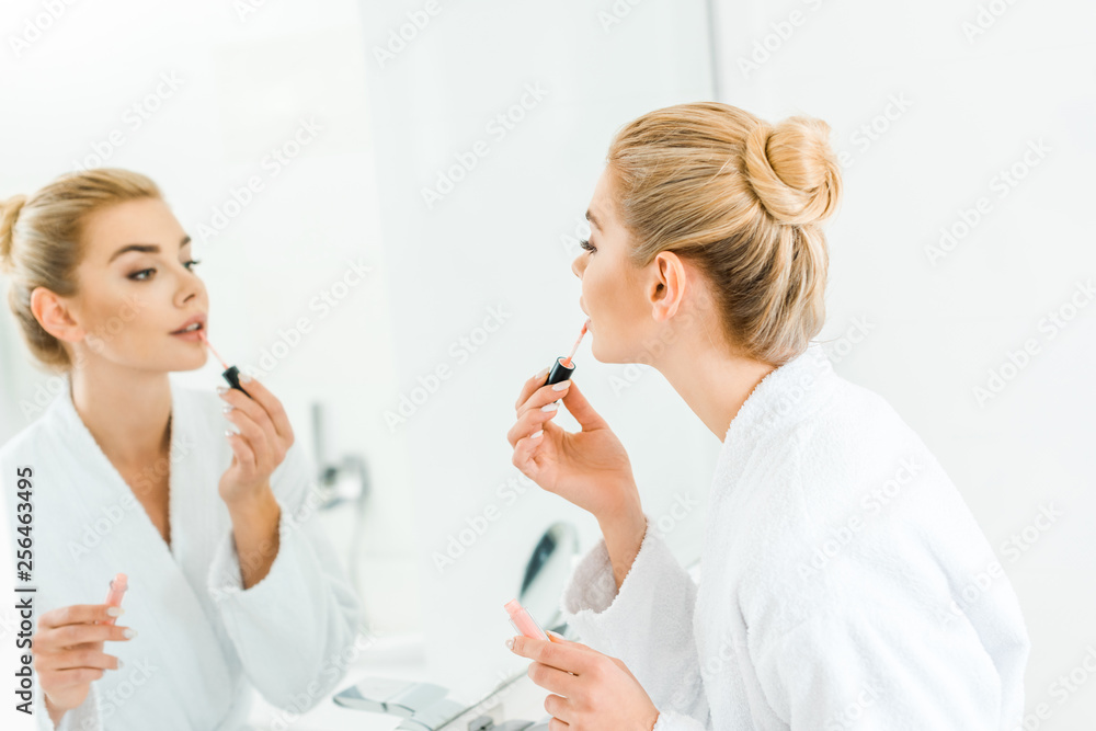 selective focus of beautiful and blonde woman in white bathrobe applying lip gloss in bathroom selective focus of beautiful and blonde woman in white bathrobe applying lip gloss in bathroom