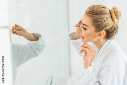 selective focus of attractive and blonde woman in white bathrobe brushing teeth with dental floss