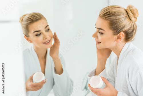 selective focus of attractive and smiling woman in white bathrobe applying face cream in bathroom