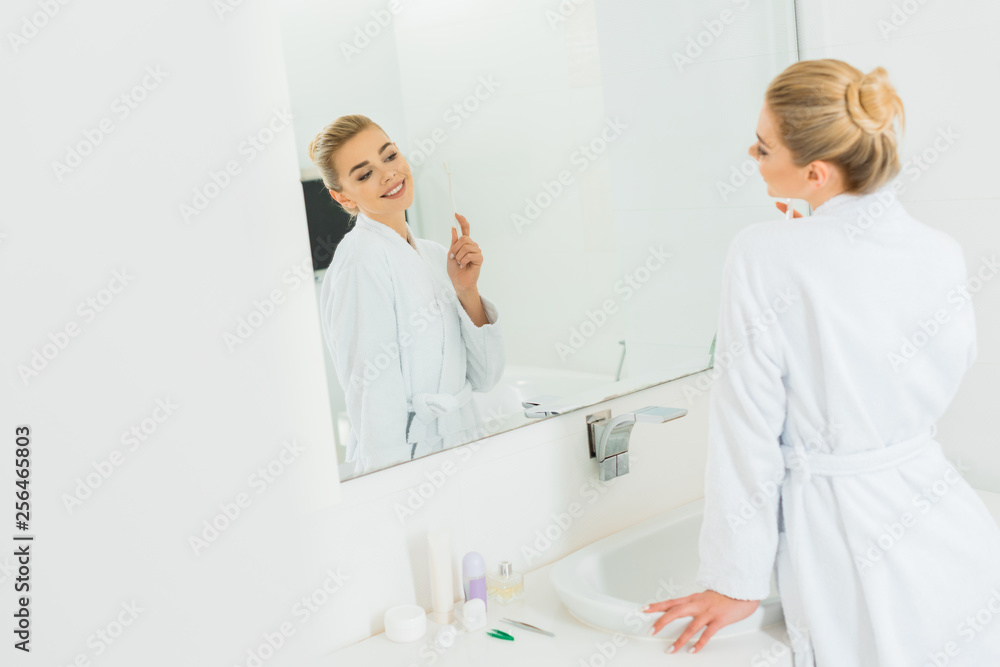 Fototapeta premium selective focus of woman in white bathrobe holding toothbrush and looking at mirror
