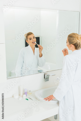 beautiful woman in white bathrobe brushing teeth and looking at mirror