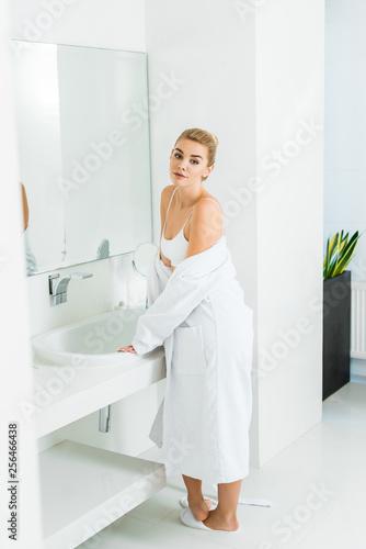 beautiful and blonde woman in bra and white bathrobe looking at camera in bathroom