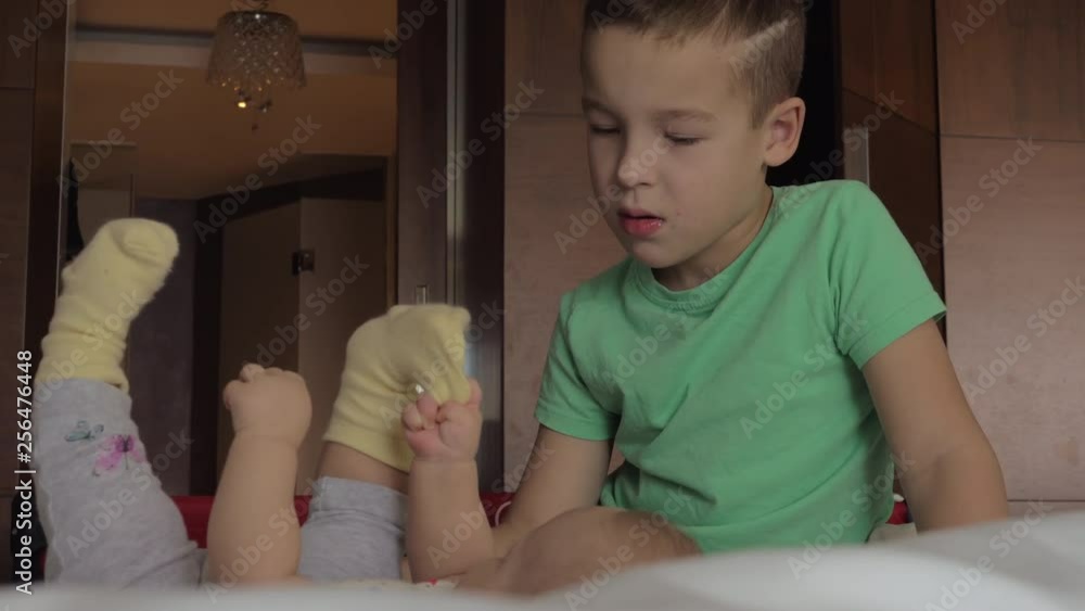 Elder brother looking at baby sister lying on the bed and laughing at her when she pulling the sock from the foot