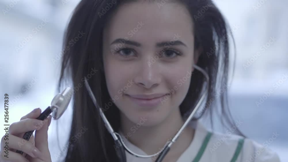 Pretty smiling female doctor face close up. Young woman with long brunette hair looking in camera holding stethoscope. Slow motion Pretty smiling female doctor face close up. Young woman with long brunette hair looking in camera holding stethoscope. Slow motion