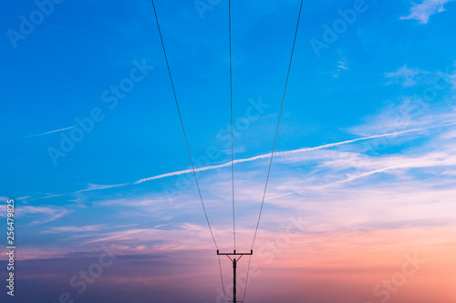 Electrical substation utility pole, state and high voltage power line silhouette with blue cloudy sky at sunset. Purple sunset view of electric tower with airplane lines on blue sky.