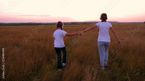 Girlfriends listening to music with headphones in field. girls hold hands and walk on thick and tall grass at sunset.