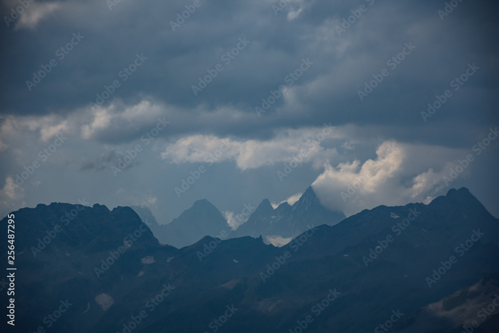 Fototapeta premium The peaks of the Caucasus Mountains in cloudy clouds and the rays of the sun through them in the outskirts of Dombai. And on the tops of snow lies.