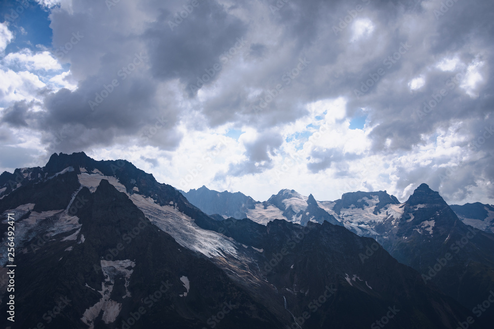 The peaks of the Caucasus Mountains in cloudy clouds and the rays of the sun through them in the outskirts of Dombai. And on the tops of snow lies. The peaks of the Caucasus Mountains in cloudy clouds and the rays of the sun through them in the outskirts of Dombai. And on the tops of snow lies.