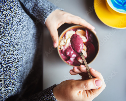 Acai smoothie, granola, seeds, fresh fruits in a wooden bowl in female hands on grey table. Eating healthy breakfast bowl. Top view photo