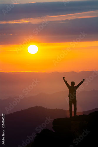 Men raise their arms in sunrise,Landscape with silhouette of a standing happy man with backpack and raised-up arms on the mountain peak on the background of cloudy sky at colorful sunset in summer.