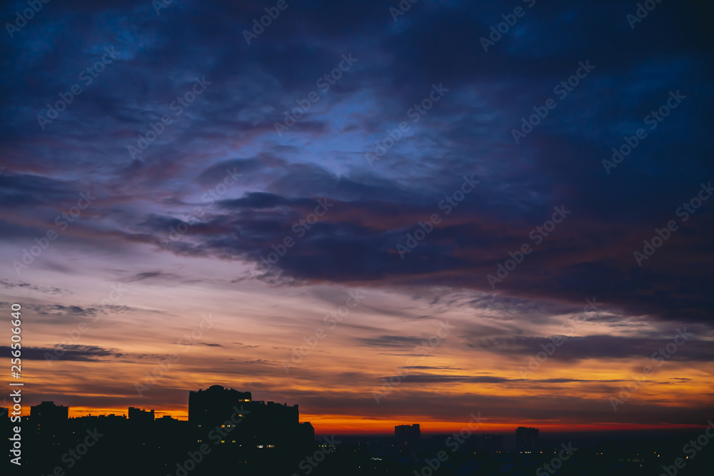 Cityscape with vivid warm dawn. Amazing dramatic blue violet cloudy sky above dark silhouettes of city buildings. Orange sunlight. Atmospheric background of sunrise in overcast weather. Copy space.