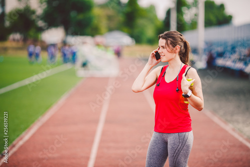 The topic is sport and health. A young Caucasian woman in training in sportswear is talking using a mobile phone, the hand is ear phone, at the sports stadium, the weather is sunny.