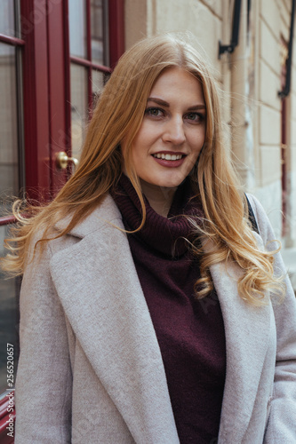 Portrait of attractive blonde woman in stylish coat looking at the camera while standing on the city street. Spring day