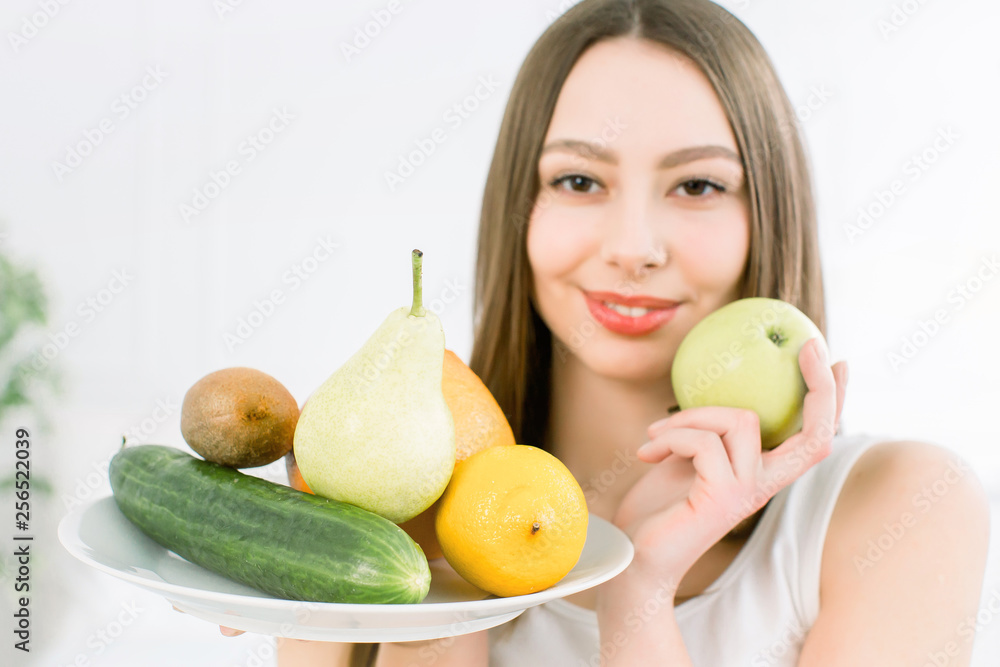 Attractive smiling young Caucasian woman holding a plate with green yellow vegetables and fruits on the light background. Healthy, beauty, good food vegetarian products. Attractive smiling young Caucasian woman holding a plate with green yellow vegetables and fruits on the light background. Healthy, beauty, good food vegetarian products.