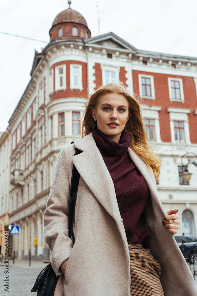 Fototapeta premium Portrait of beautiful young blonde woman in casual coat looking at the camera on the city street. Beautiful architecture building on background