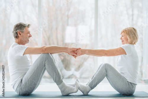 Sports and healthy lifestyle.Sporty middle aged couple sitting on the mat in the white fitness gym and doing stretching while looking each other and smiling. Healthy joints and back. Support trainer. photo