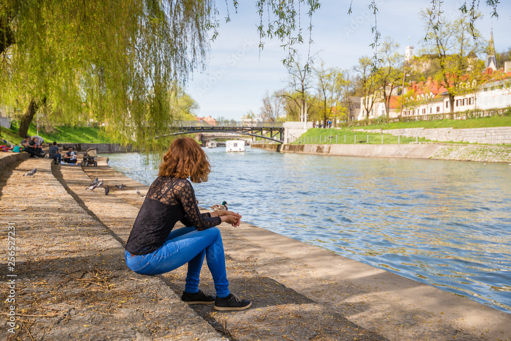 Young woman is sittng on the banks of Ljubljanica river at Trnovo "beach"  admiring the beautiful Ljubljana old town in Ljubljana, Slovenia. Stock  Photo | Adobe Stock