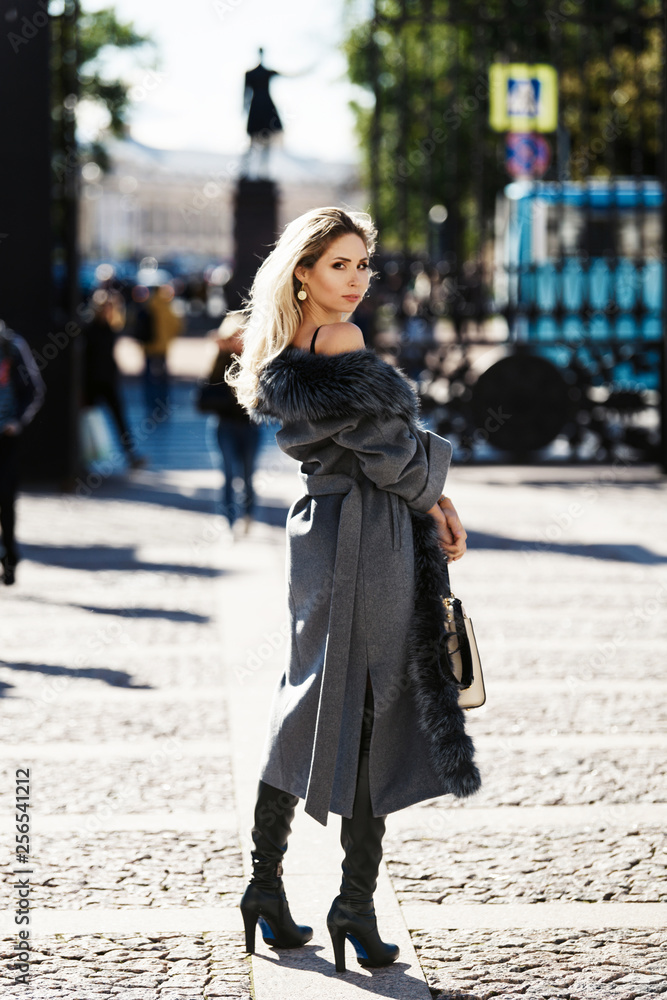 Beautiful young woman in fur coat, smiling, posing with luxury bags and accesories while standing and walking outdoors. Female fashion city lifestyle outfit spring or autumn shopping concept