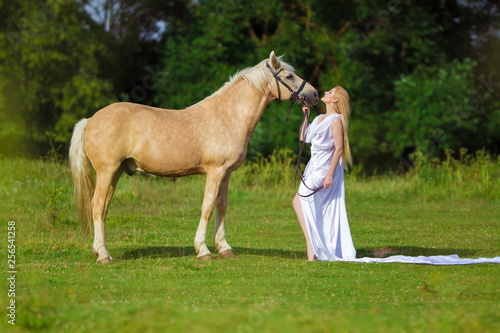 Young rider woman blonde with long hair in a white dress with a train posing and jumping on a palamino horse against a field and forest background photo