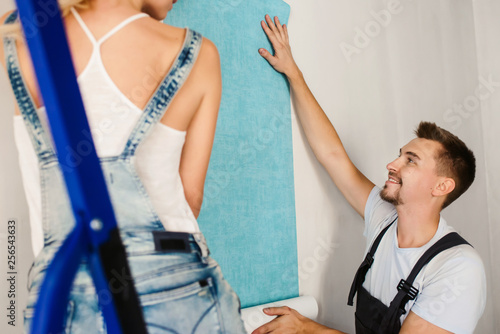 Funny couple, woman and her husband wallpapering the walls using a metal ladder. Home renovation and moving to new house concept