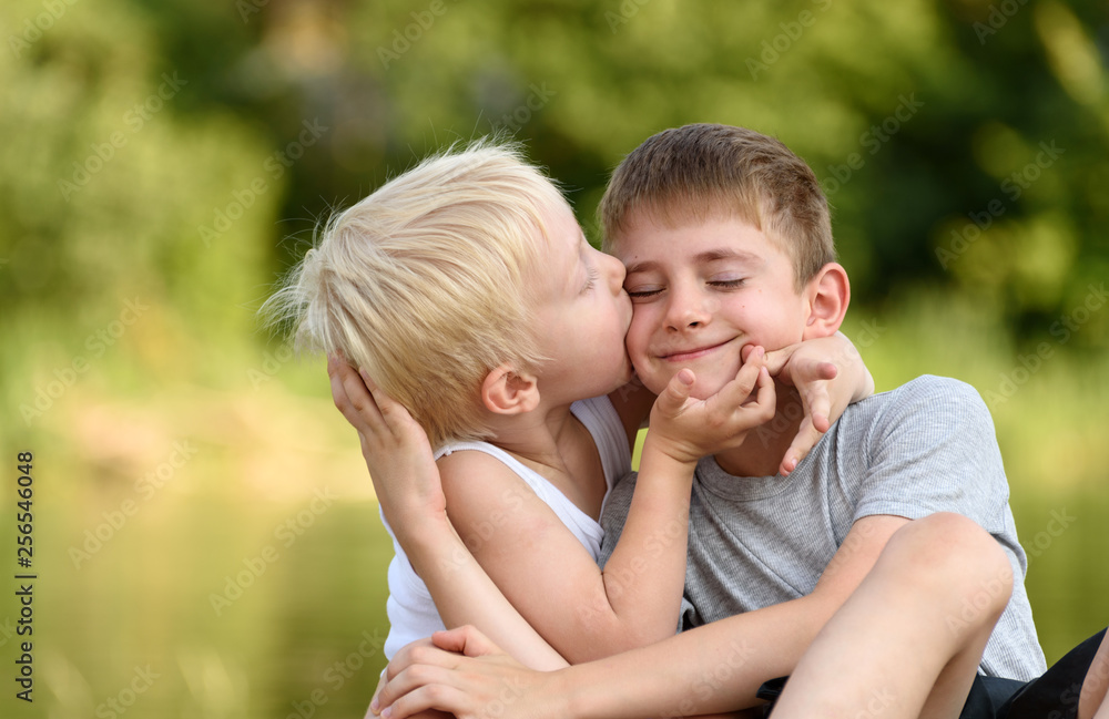 Two little brothers are sitting outdoors. One kisses the other on the cheek. Blurred green trees in the distance. Concept of friendship and fraternity. Two little brothers are sitting outdoors. One kisses the other on the cheek. Blurred green trees in the distance. Concept of friendship and fraternity.