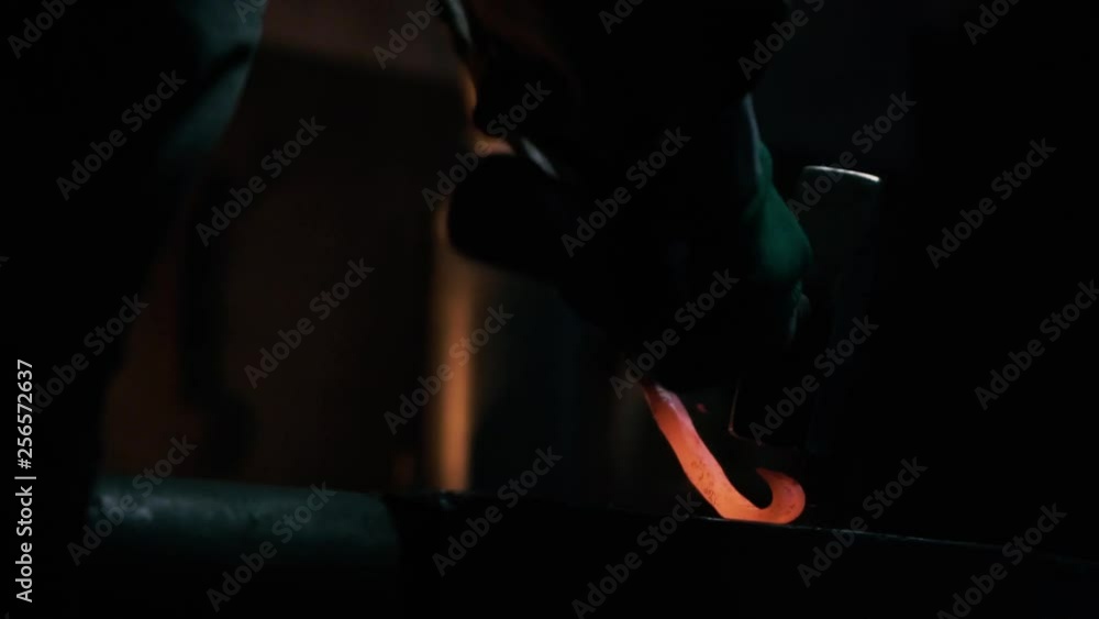 Blacksmith swings metal hammer forging hook from steel bar in dark workshop with fireplace in background. Blacksmith swings metal hammer forging hook from steel bar in dark workshop with fireplace in background.