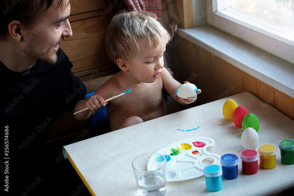 Fototapeta premium Father and his toddler blond son dyeing Easter eggs together at home, they laugh and have fun together