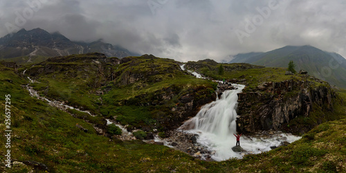 man in red stands under waterfall on black rock in mountains on cloudy rainy day.. Wide panorama