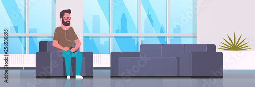 young man sitting armchair listening to music on smartphone with headphones bearded guy resting at home modern living room interior flat horizontal banner full length