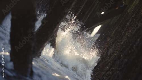 Waves crashing under a pier, old wooden pillings with sunbeams coming through. Storm. Medium shot dutch angle, angled. photo