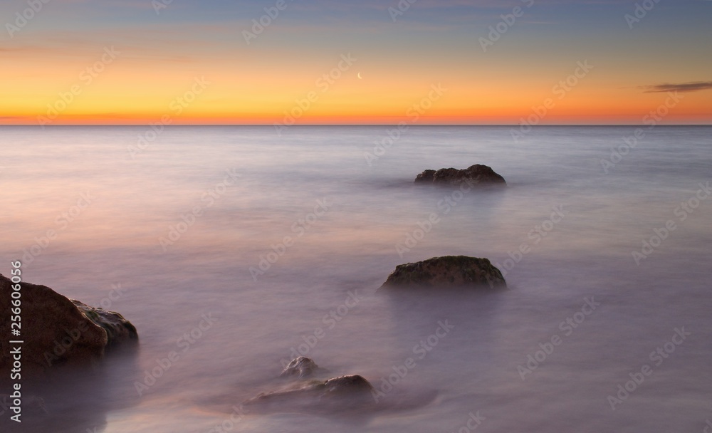 Crescent moon at sunrise with beautiful red sky, calm sea with rocks in foreground, port nou, cala bona, mallorca, spain.