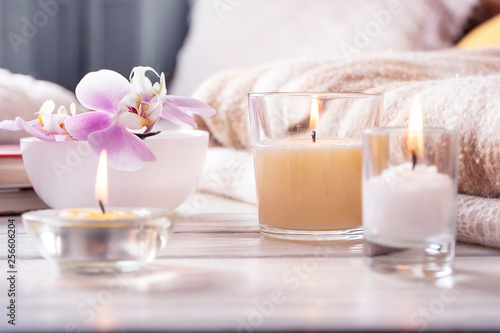 Composition of three candles, orchid in vase on white wooden tray in front of bed. Home interior detailes close up.
